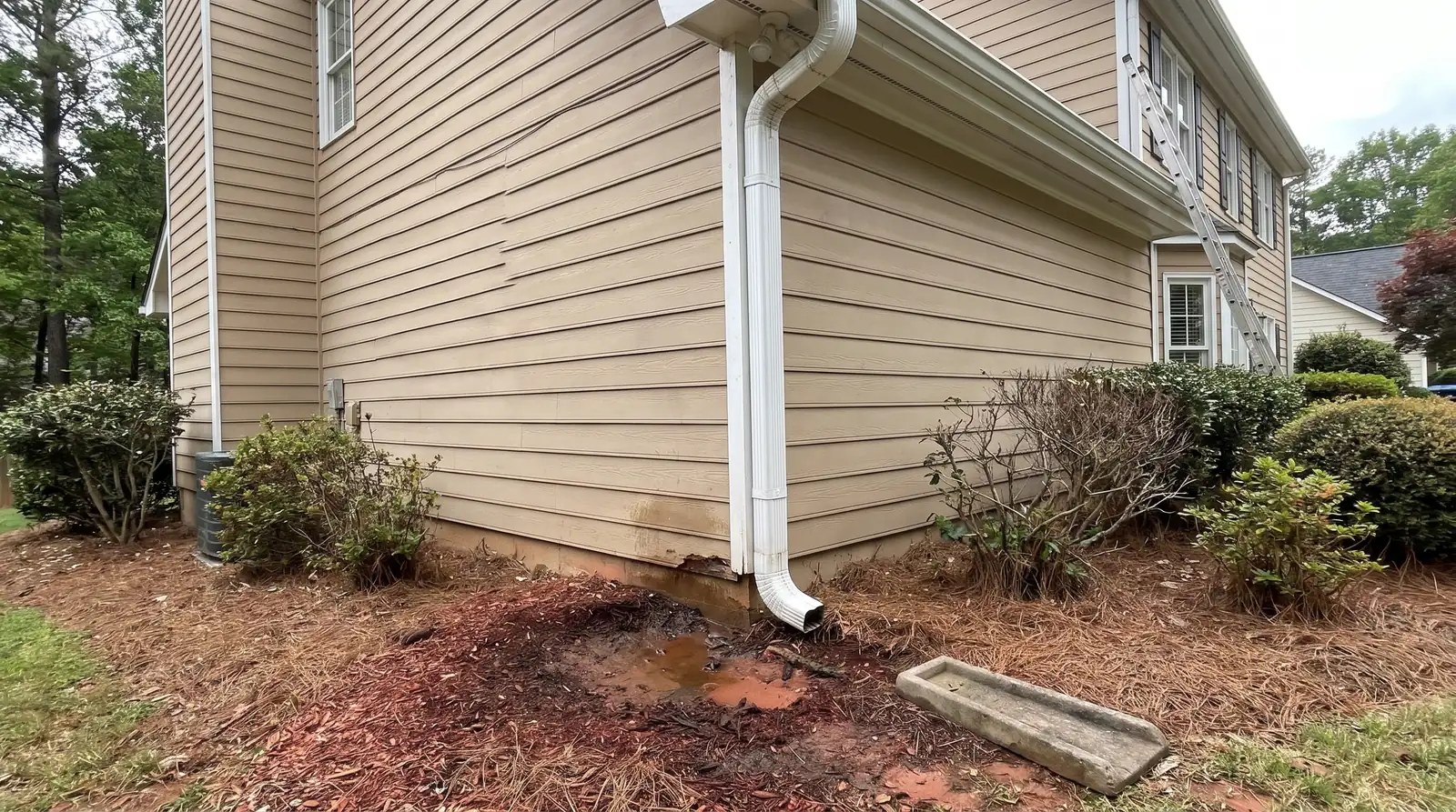 Downspout emptying against a house's foundation into washed-out red clay, its splash block left aside